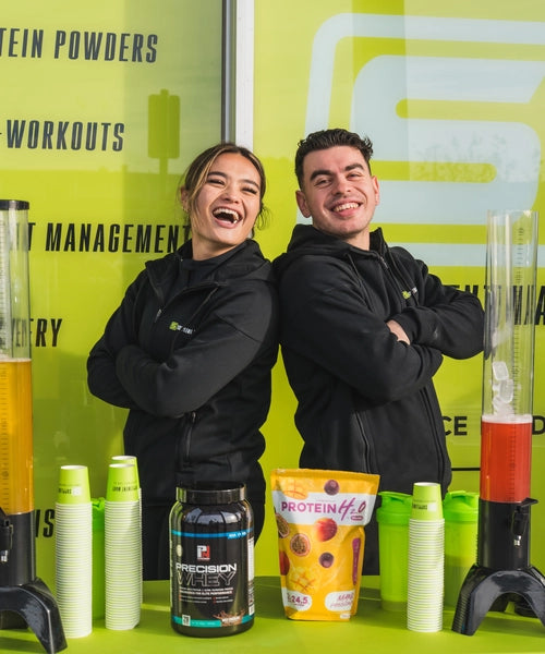 Supplement Mart staff standing in front of a Supplement Mart building with shakers and samples on the table in front of them as they are smiling and happy